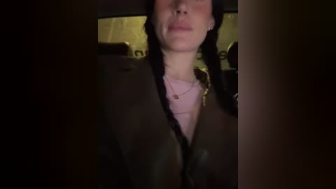 Video of a person in a dimly lit car, wearing a pink shirt, black jacket, and a necklace, with their face partially obscured by darkness.