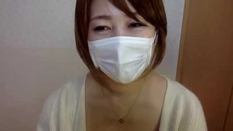 A video of an Asian woman with short brown hair, wearing a white surgical mask, a white cardigan, and a gold necklace. She has a neutral expression. The background is a plain beige wall and wooden door.