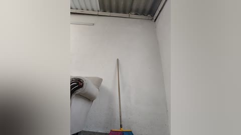 Video of a small, empty room with white walls, a metal ceiling, and a white roll of paper on the floor. A single long pole leans against the wall, and a colorful object is partially visible in the bottom right corner.