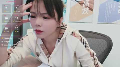 Video of a young Asian woman with straight black hair, wearing a white and brown plaid jacket, sitting at a desk, looking tired with her hand on her forehead.