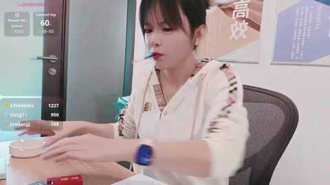 A video of an Asian woman in a white shirt, blue nails, and a smartwatch, sitting at a desk in an office, looking at her phone.
