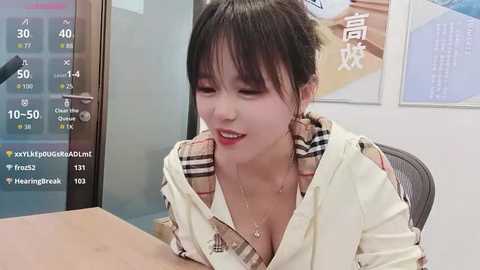 Video of an Asian woman with medium-length black hair, fair skin, wearing a white shirt with a plaid collar, sitting at a table, smiling. Background includes a glass door and Chinese text.