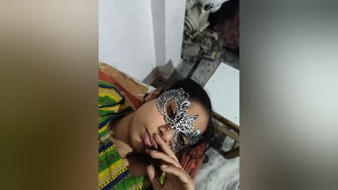 Video of a young woman with light brown skin, wearing a colorful, patterned sari and a detailed, ornate masquerade mask. She has dark hair and is seated in a room with white walls and a tiled floor, with a large black-and-white framed portrait behind her.