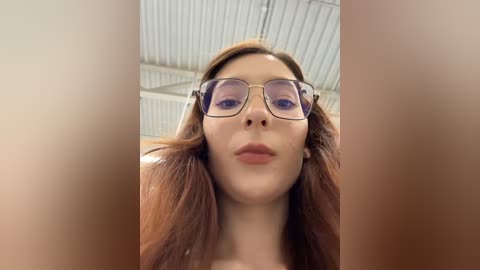 A close-up video of a young woman with fair skin, brown hair, and wearing large, rectangular glasses, taken from a low angle, showing a portion of her upper body. The background features a corrugated metal roof.