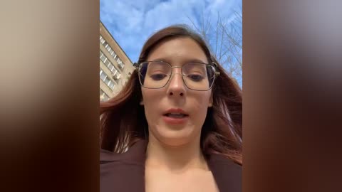 A video of a young woman with fair skin, brown hair, and rectangular glasses, taken from a low angle, with a blurry, wooden background and a building visible in the background.