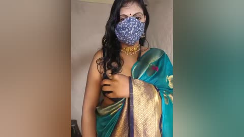 Video of a South Asian woman with medium-dark skin, wearing a blue and gold saree, blue mask, and gold necklace, covering her chest with her right hand. Background shows beige and green walls.