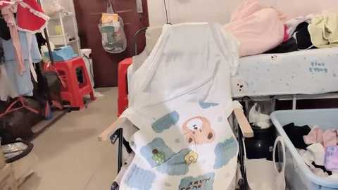 Video of a cluttered hospital room with a baby stroller covered in cartoon-themed fabric, surrounded by medical equipment, toys, and a red plastic chair.