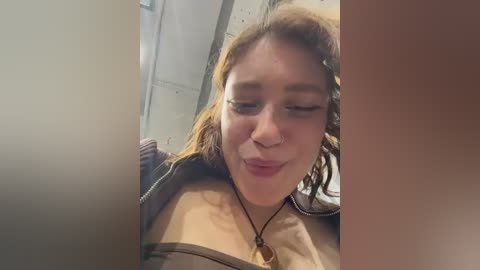 Video of a young woman with light skin and brown hair, wearing a brown jacket, making a funny face, with a blurred background.