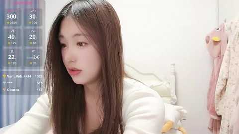 Video of an Asian woman with long, straight brown hair, wearing a white shirt, sitting in a cozy bedroom with a pink teddy bear, a pink towel hanging, and a temperature display.