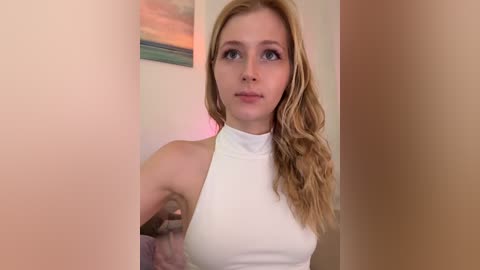 A video of a young Caucasian woman with long, wavy blonde hair, wearing a sleeveless white top, standing in a brightly lit room with a framed painting on the wall.