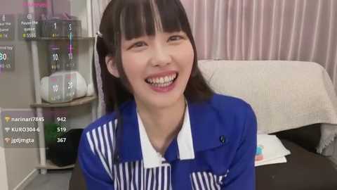 Video of an East Asian woman with straight black hair, wearing a blue and white striped shirt, smiling, sitting in a medical office with a beige wall and white curtains.