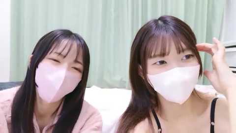 Media: Video of two young East Asian women with long black hair, wearing white face masks, sitting on a bed, one adjusting her mask. Pale green curtains in the background.