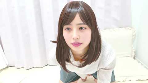 Video of an East Asian woman with shoulder-length straight black hair, wearing a white knitted sweater and blue denim skirt, sitting on a white cushioned chair with a white curtain in the background.