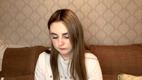 Video of a young woman with straight, light brown hair, wearing a white T-shirt, sitting on a brown couch against a wallpapered wall.
