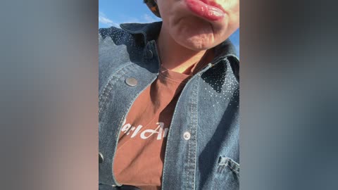 Video of a young person with light skin, wearing a denim jacket over a brown t-shirt with \"Burger King\" text. They stick their tongue out playfully, under bright sunlight.