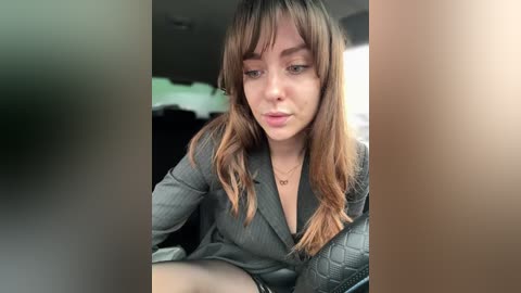 Video of a young woman with fair skin and shoulder-length brown hair, wearing a gray pinstripe blazer, seated in a car. She looks contemplative, with a neutral expression. The background is blurred, featuring a dimly lit interior.