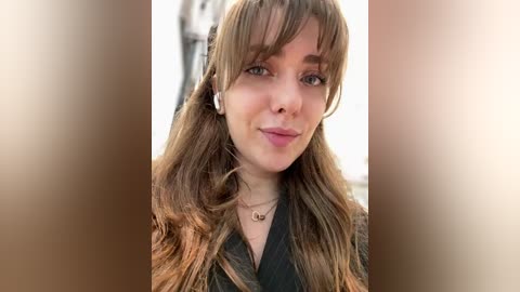 A video of a young woman with long, wavy brown hair, wearing a black jacket and white earphones, smiling subtly. The background is blurred, with a warm, neutral tone.