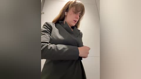 Video of a Caucasian woman with light brown hair and bangs, wearing a dark gray pinstripe suit, standing in a white hallway, arms crossed, looking serious.