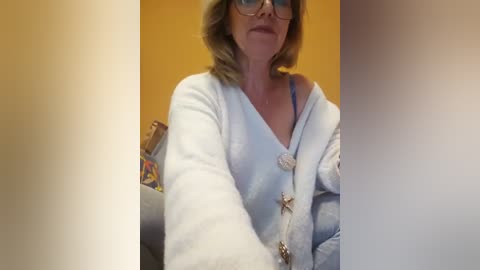 Video of a middle-aged woman with shoulder-length blonde hair, wearing glasses and a blue robe, sitting in a cozy room with yellow walls, holding a bag.