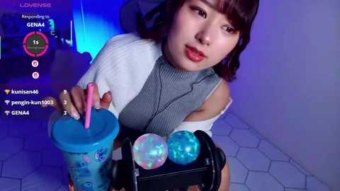 A video of an Asian woman with straight, shoulder-length brown hair, wearing a grey ribbed top and holding a blue cup with a pink straw. She's in a room with a blue and purple light.