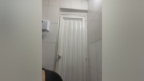 Video of a small, narrow bathroom with white tiled walls, a white door with vertical blinds, a white toilet paper dispenser, and a partially visible person in black clothing.