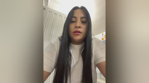 Video of a young woman with long, straight black hair, wearing a white T-shirt, standing in a beige-walled bathroom. Background features a white cabinet and toiletries.