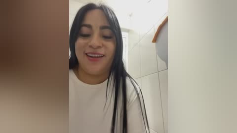 Video of a Latina woman with long black hair, wearing a white shirt, smiling in a bathroom with tiled walls and a wooden shelf.