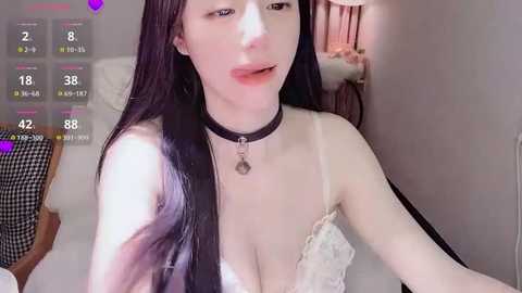Video of a fair-skinned, slim Asian woman with long black hair, wearing a black choker and a white lace bra, posing seductively in a bedroom.