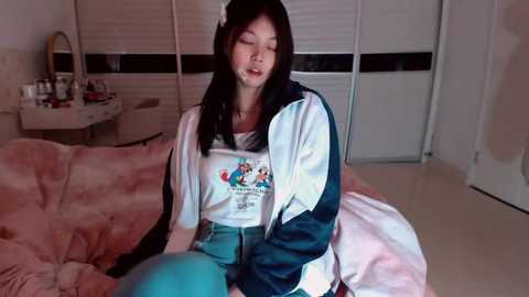 A video of an Asian woman with long black hair, wearing a white graphic t-shirt and blue jeans, sitting on a pink bed, eyes closed, in a dimly lit bedroom.