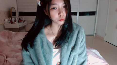 A video of an Asian woman with long black hair, wearing a teal fur robe, posing indoors on a bed with pink sheets. The background includes a vanity with makeup, a mirror, and white walls with blinds.