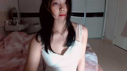 Media: Video of a slender Asian woman with long black hair, wearing a white lace top, sitting on a pink bed in a dimly lit bedroom with closed white blinds and a vanity table.