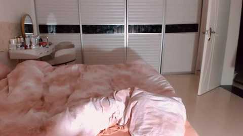 Video of a messy bedroom with a pink, fluffy comforter, a vanity table, a round mirror, a closed white door, and a sliding white closet door.