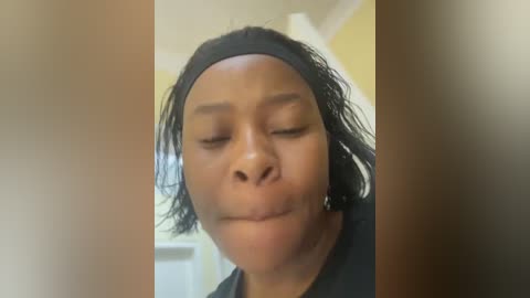 A close-up video of a Black woman with medium brown skin and shoulder-length, wavy black hair, wearing a black headband, smiling with her eyes closed. She has a slight dimple on her right cheek.