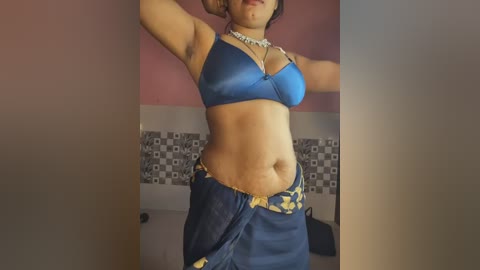 Video of a woman with medium-dark skin, wearing a blue bra and a blue skirt with gold embroidery, posing in a room with pink walls and a tiled backsplash.