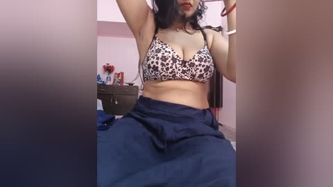 Media: Video of a woman with medium skin tone, wearing a leopard print bra and blue skirt, sitting on a bed in a dimly lit room with a nightstand and various items in the background.