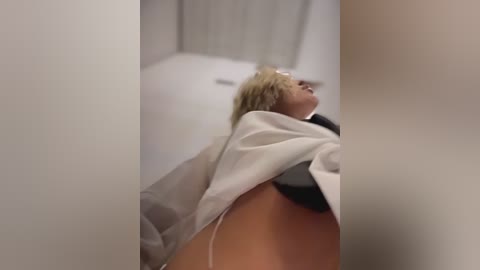 Video of a blonde woman lying on a white bed, wearing a white robe, with a black bra visible, in a dimly lit, white-walled room.