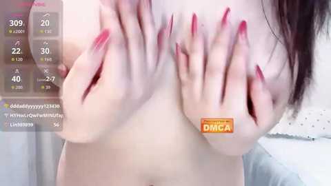 Video of a topless woman with fair skin, large breasts, and red manicured nails. She is pressing her breasts together, partially covering them. Background shows a white wall and a digital clock displaying 39 degrees.