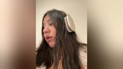 Video of a woman with long, dark hair wearing large white headphones, looking off to the side with a neutral expression. Background is blurred.