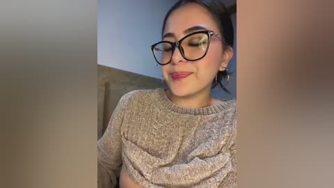 Media: Video of a young woman with medium skin tone, dark hair, and black-framed glasses, wearing a beige knit sweater. She smiles softly, standing against a blurred beige wall.