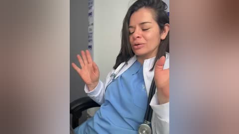 A video of a young woman with fair skin and dark hair, dressed in blue medical scrubs and a white shirt, holding a stethoscope and gesturing with her right hand.