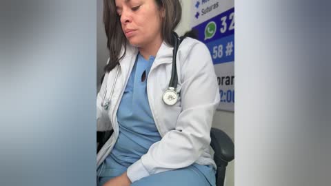 Video of a female nurse with long brown hair, wearing a white lab coat over light blue scrubs, sitting in a hospital room with a stethoscope around her neck, and a sign with medical information in the background.