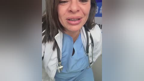 Video of a middle-aged woman with medium-length brown hair, wearing a white lab coat over light blue scrubs, smiling, with a stethoscope around her neck, in a medical setting.