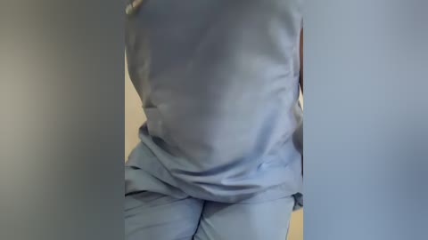 Video of a person wearing a light blue hospital gown, standing in a dimly lit room with beige walls. The gown is slightly wrinkled, and the person's arms are not visible.