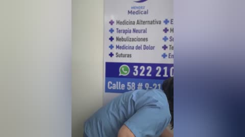 A video shows a person wearing a light blue shirt sitting on a white medical office chair, with a medical directory poster in the background listing services.