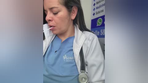 Video of a Latina healthcare worker in blue scrubs and a white lab coat, using a stethoscope, with blurred background showing medical posters.