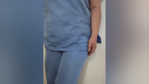 Video of a person wearing light blue scrubs, standing against a plain white wall. The person's left arm is bent, hand resting on the wall. The background is minimalistic, focusing on the subject's attire and posture.