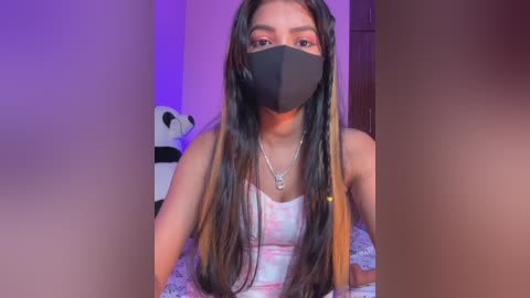 Video of a young woman with long, dark hair, wearing a black face mask, white tank top, and silver necklace, seated indoors with a purple and pink background, featuring a stuffed panda bear.