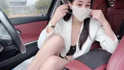 A video of an Asian woman with long black hair, wearing a white blazer, black lace bra, and a face mask, sitting in a red leather car seat.