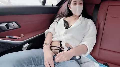 Video of a young woman with long dark hair, wearing a white button-up shirt and black lace bra, sitting in a luxury car with red leather seats, wearing a face mask.