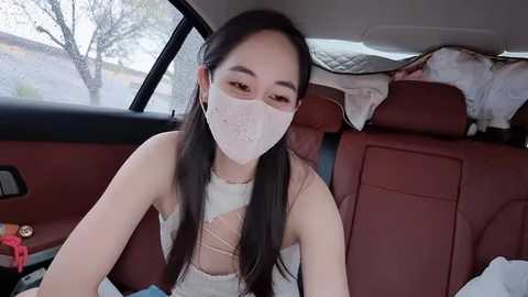 Video of a young Asian woman with long black hair, wearing a white lace face mask, sitting in the back seat of a car.